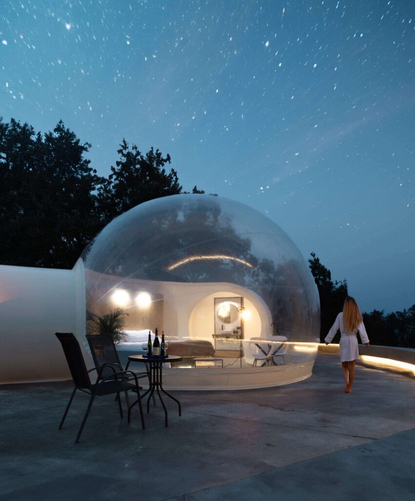 Burbujas Balú transparente iluminada en la noche bajo un cielo estrellado, con cama doble, terraza con mesa y sillas, y una mujer en albornoz caminando hacia el alojamiento