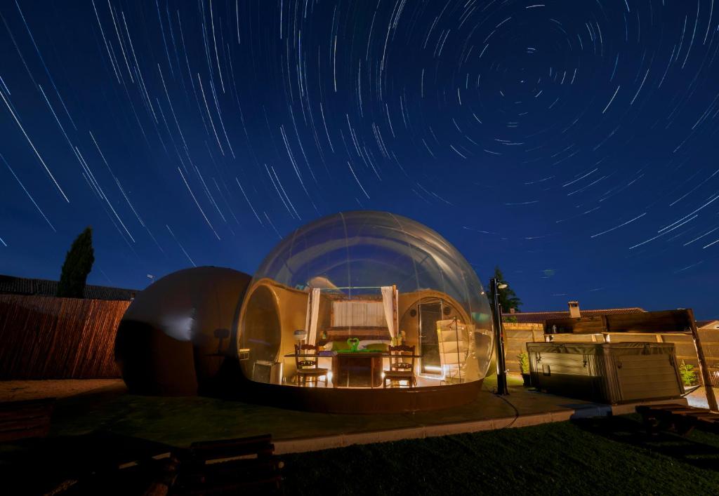 Burbuja transparente iluminada por la noche con cama con dosel y jacuzzi exterior, bajo un cielo estrellado con efecto de estelas de estrellas
