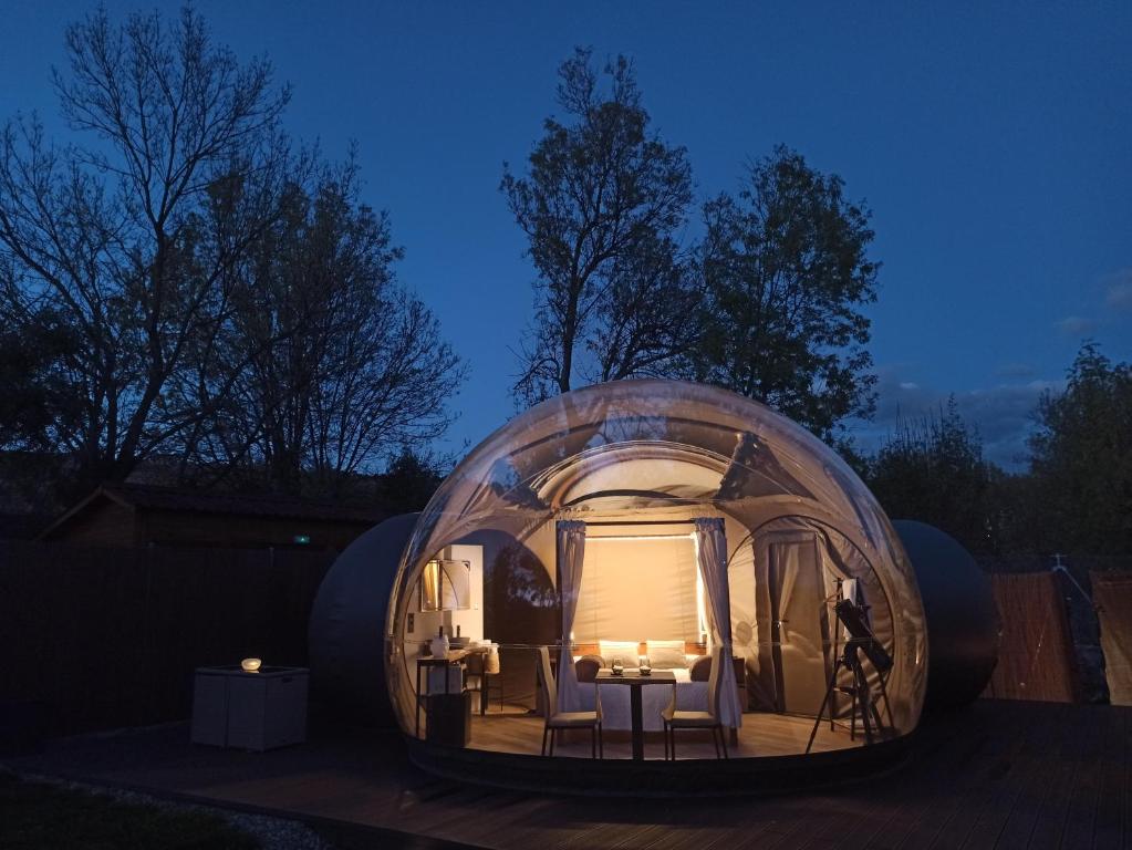 Imagen de una burbuja transparente iluminada desde el interior durante el atardecer o la noche, ubicada en una plataforma de madera en medio de un entorno natural con árboles; en su interior se observa una cama con dosel, una mesa con sillas, un telescopio y detalles decorativos que transmiten una atmósfera acogedora y romántica