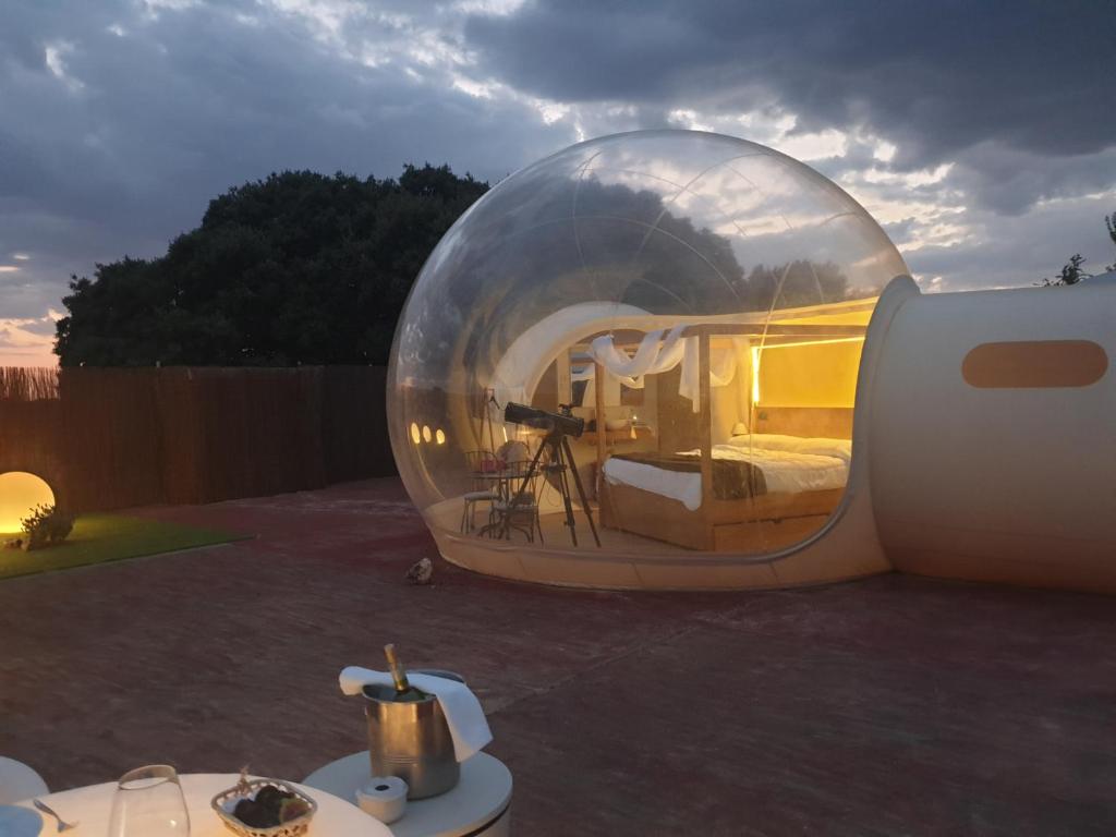 Burbuja transparente iluminada al anochecer con cama con dosel, telescopio orientado hacia el cielo y mesa exterior preparada para una cena romántica