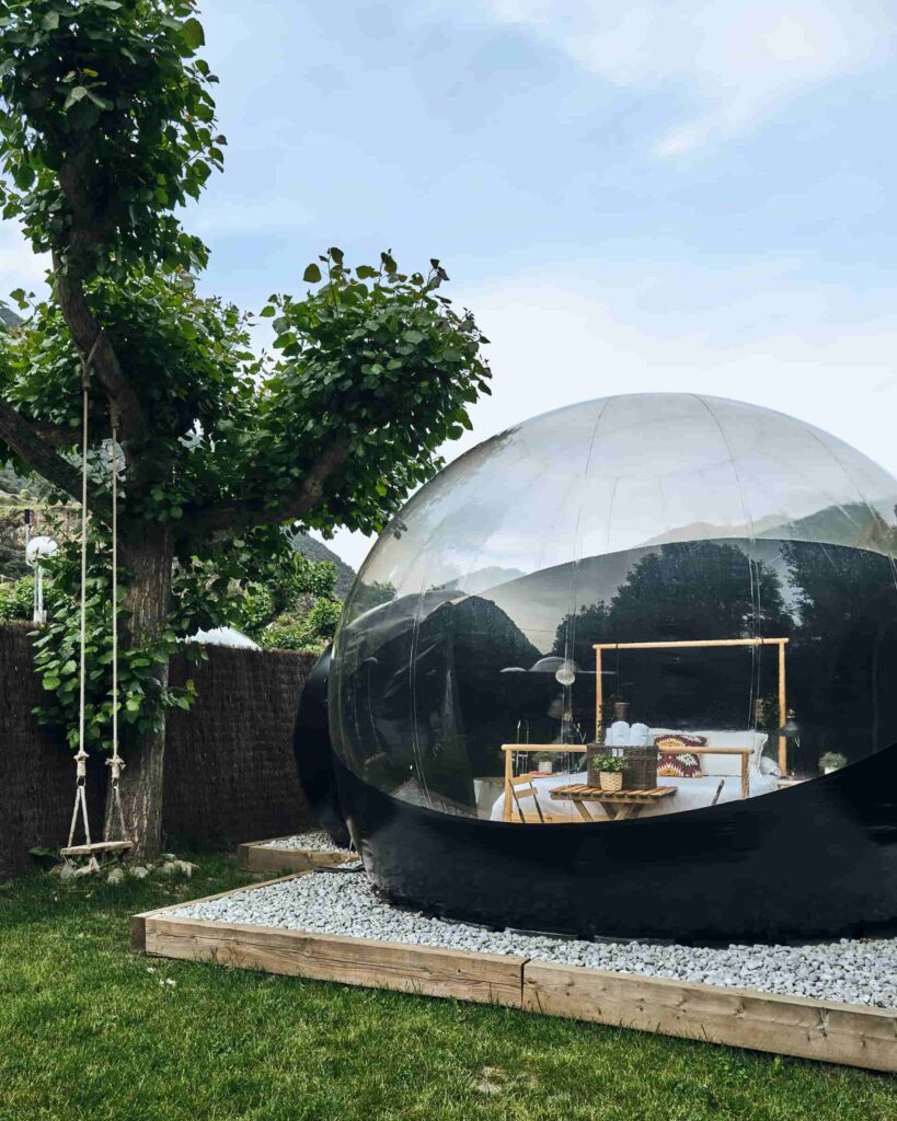 Burbuja de glamping negra con interior visible, ubicada en un jardín con césped, árboles y un columpio de cuerda
