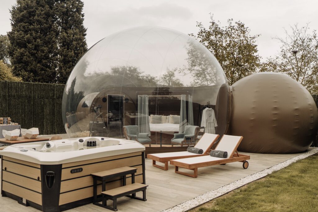 Burbuja transparente de lujo con cama doble y zona de estar en su interior, acompañada de dos tumbonas y un jacuzzi exterior de madera, rodeada de árboles y vegetación