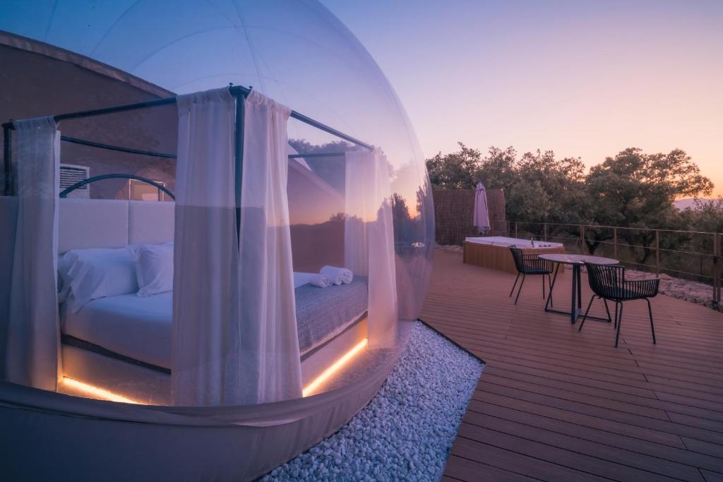 Habitación burbuja con cama con dosel iluminada al atardecer, en una terraza de madera con jacuzzi y muebles de exterior, rodeada de naturaleza
