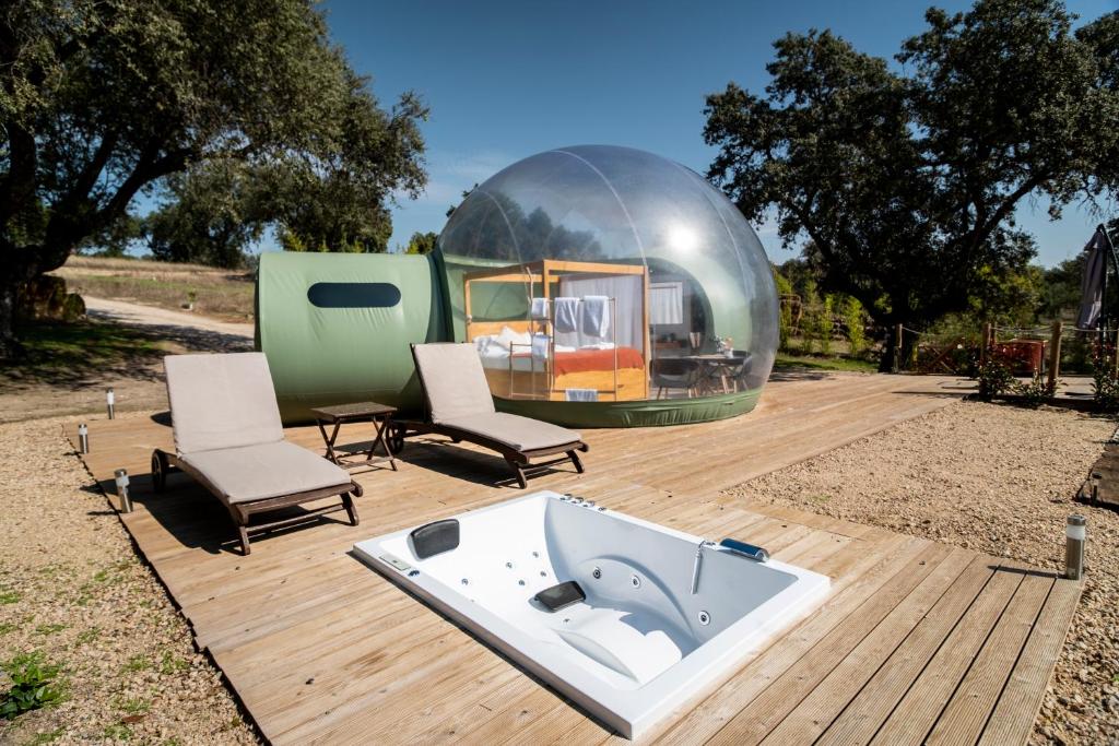 Burbuja transparente de glamping con cama en su interior, acompañada de tumbonas y jacuzzi exterior sobre una plataforma de madera, rodeada de árboles y naturaleza en un día soleado