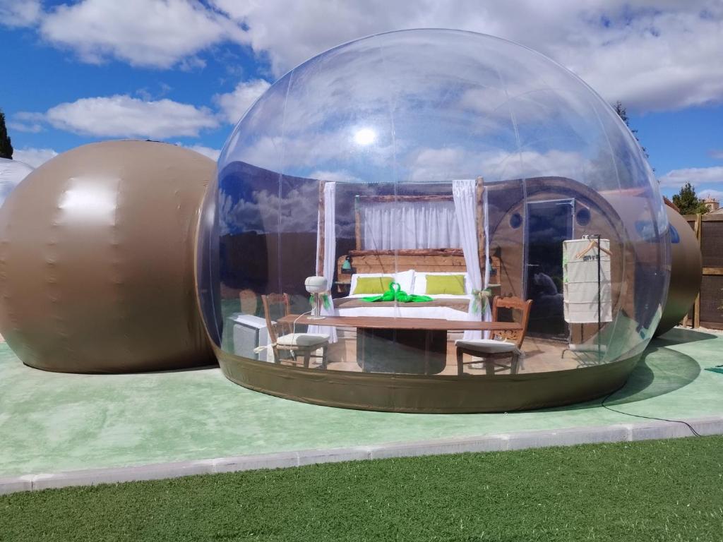 Burbuja transparente con cama, cortinas blancas y mobiliario interior, ubicada sobre una plataforma junto a césped artificial, bajo un cielo parcialmente nublado