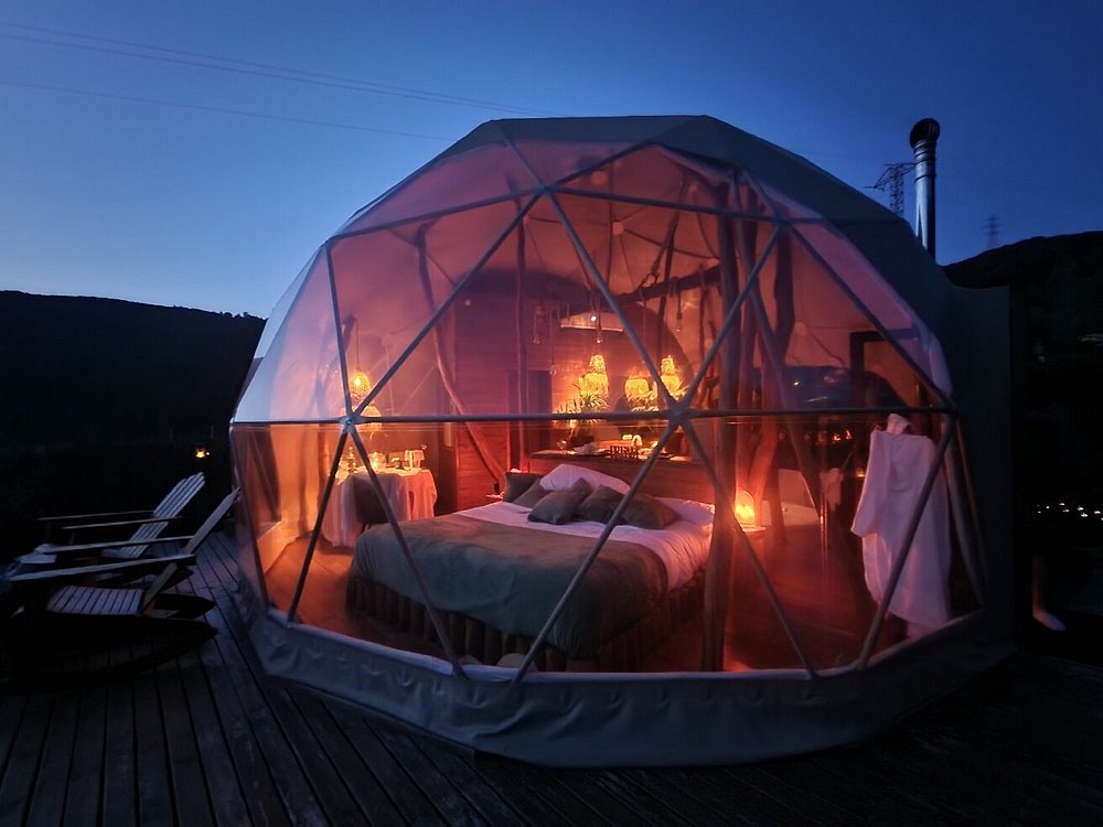 Cúpula de lujo iluminada por la noche en medio del bosque, con cama doble, decoración acogedora y vistas panorámicas desde una estructura geodésica transparente