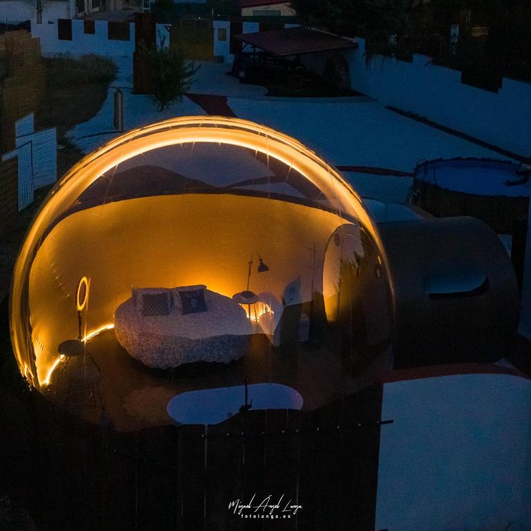 Burbuja transparente iluminada por dentro con cama redonda, lámparas y decoración acogedora, ubicada en un espacio exterior rodeado de muros y vegetación, durante la noche