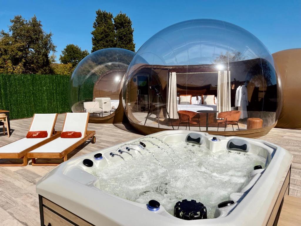 Burbuja transparente de lujo con cama doble y cortinas, acompañada de jacuzzi exterior en funcionamiento y dos tumbonas con cojines, en una terraza de madera al aire libre bajo cielo despejado
