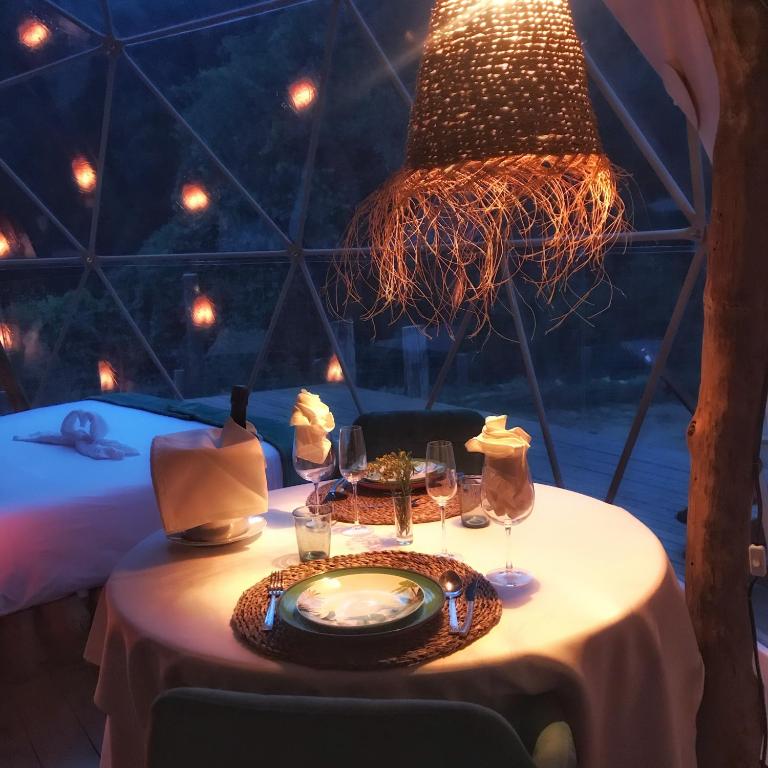 Mesa elegantemente preparada para una cena romántica dentro de una burbuja con vistas al bosque, iluminada con una lámpara rústica de fibras naturales