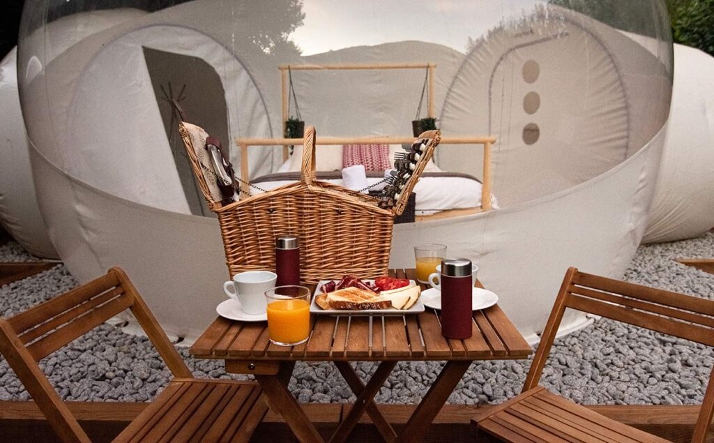 Mesa de desayuno con cesta de picnic frente a burbuja de glamping con cama doble al fondo