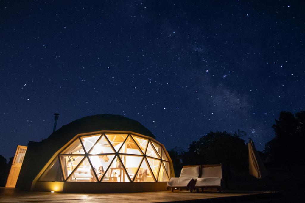 Domo geodésico iluminado por dentro durante la noche bajo un cielo estrellado, con terraza de madera y tumbonas en el exterior