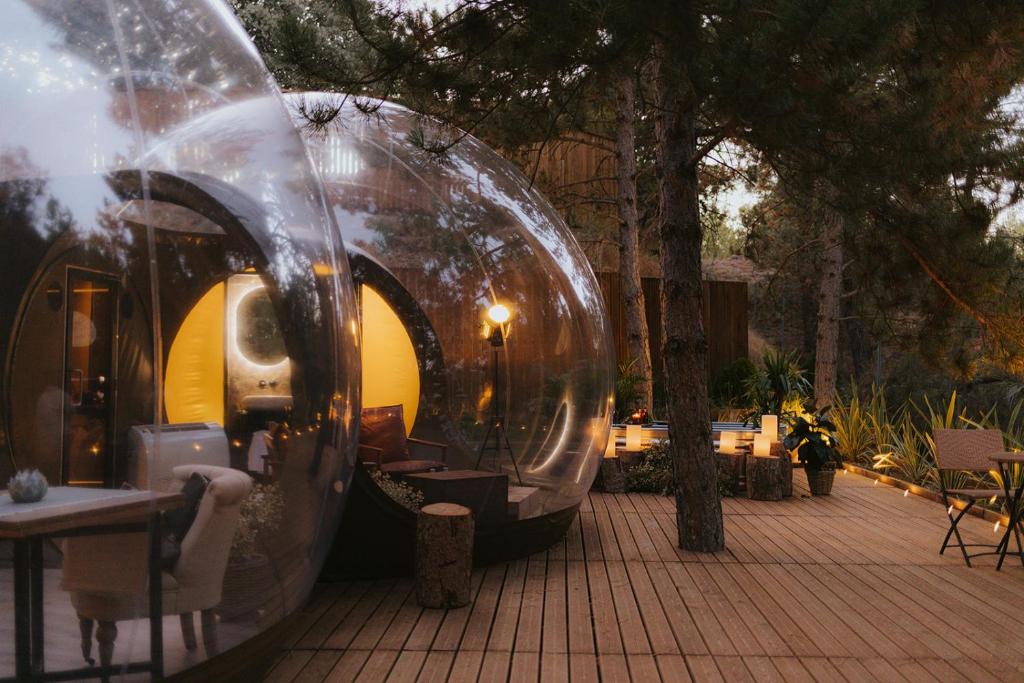 Suite de hotel burbuja de lujo, rodeada de árboles, con terraza de madera, iluminación ambiental y zona de estar al aire libre por la noche