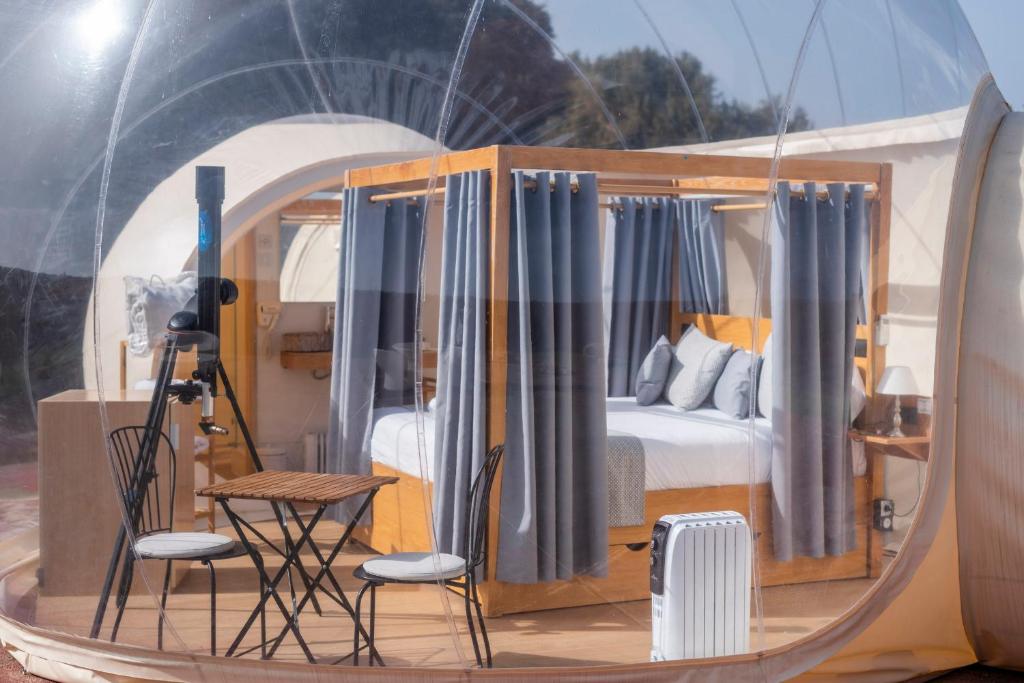 Interior de un hotel burbuja con cama con dosel, cortinas grises, telescopio, mesa exterior y calefactor portátil en un día soleado