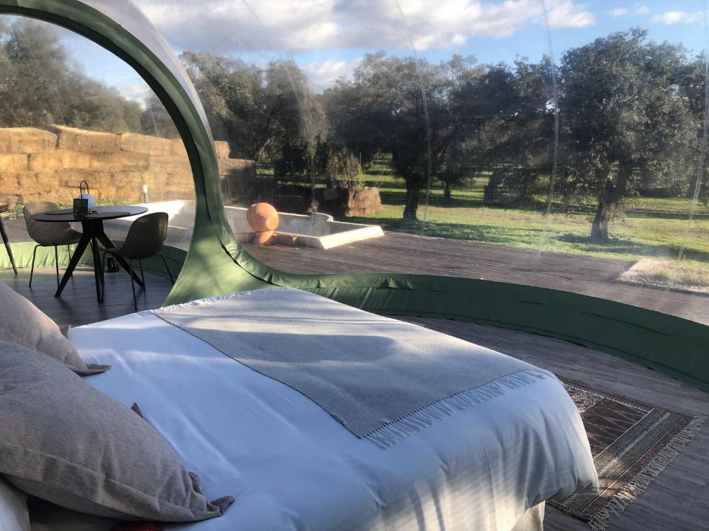Interior de una burbuja con cama doble y zona de comedor, con vistas panorámicas a un campo abierto con árboles y pacas de heno al fondo