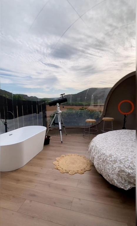 Interior de burbuja panorámica con bañera blanca independiente, cama doble, telescopio y vistas abiertas a las montañas bajo un cielo parcialmente nublado