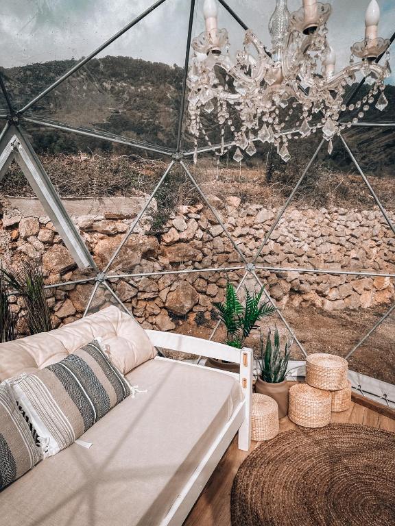 Interior de una burbuja geodésica decorada con estilo rústico chic, sofá con cojines, alfombra de yute, lámpara de araña y vistas a un paisaje montañoso con muros de piedra
