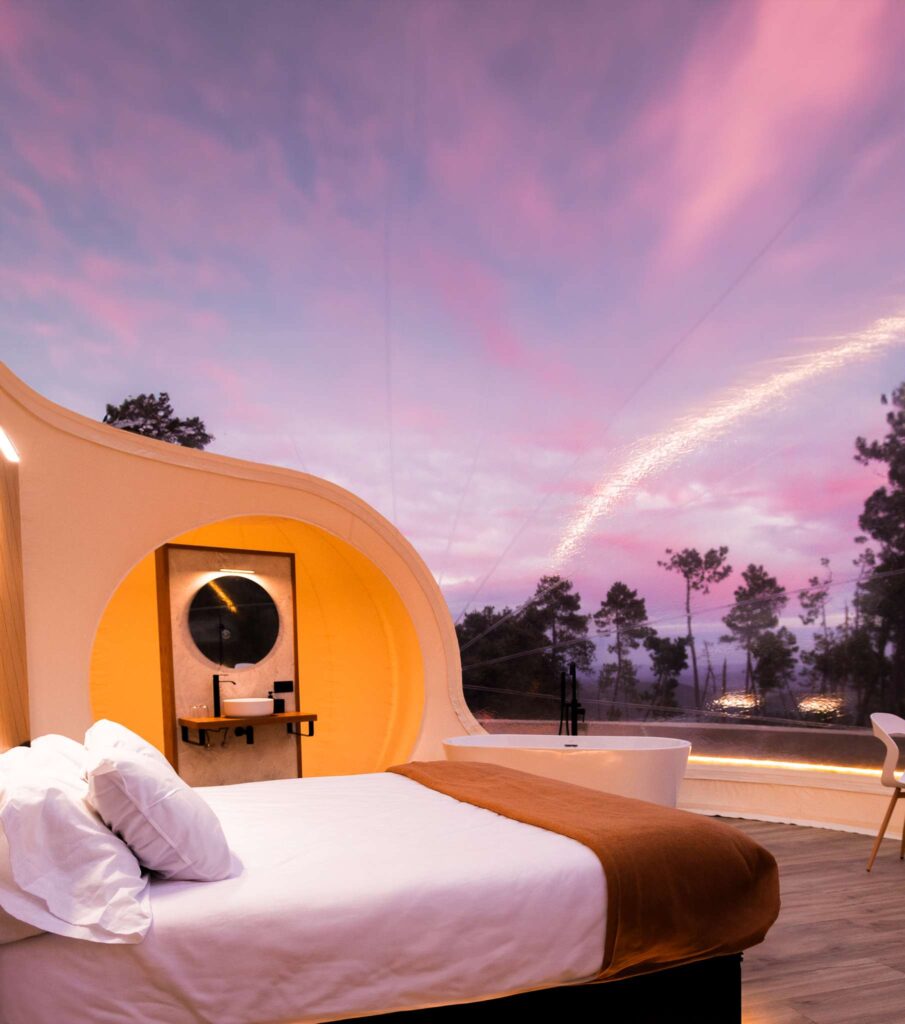 Interior de burbuja de lujo con cama doble, baño integrado y bañera frente a un gran ventanal con vistas al bosque al atardecer, bajo un cielo rosado y violeta.