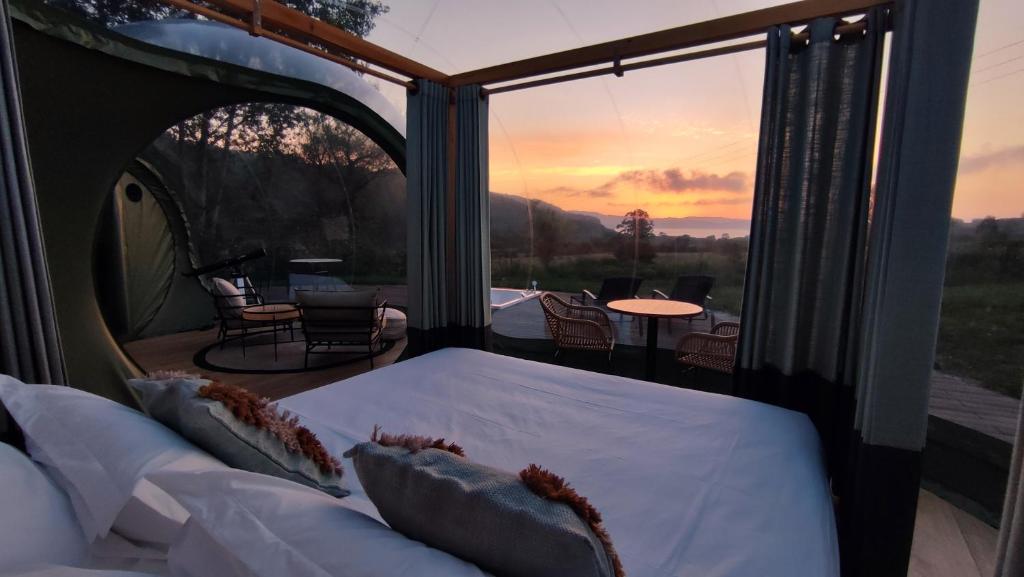Vista desde el interior de una burbuja glamping con cama doble hacia una terraza al aire libre, con mobiliario exterior y un espectacular atardecer sobre el paisaje montañoso