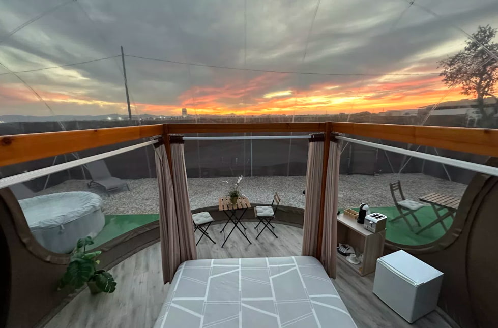 Vista desde el interior de una burbuja transparente con cama doble, terraza privada y jacuzzi exterior al atardecer