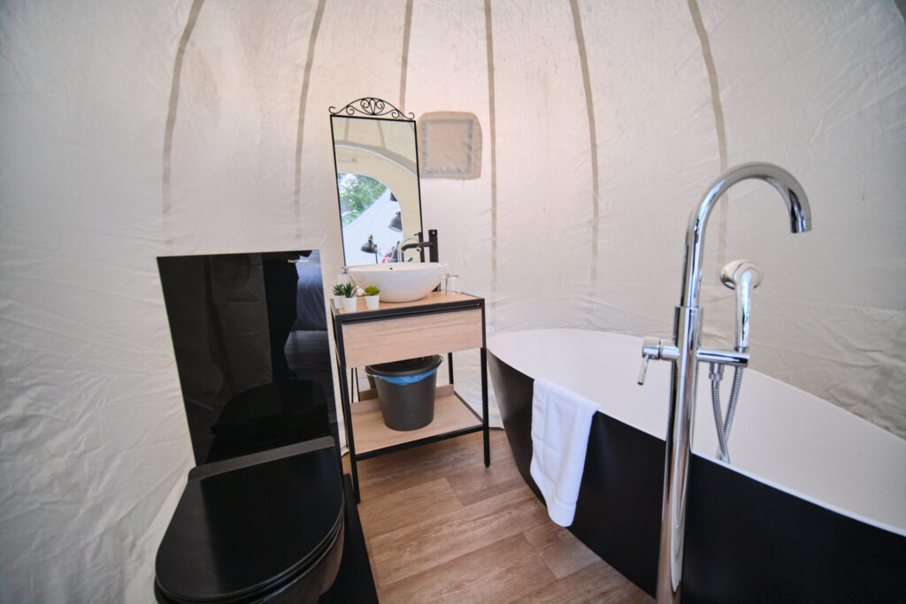 Baño interior de burbuja glamping con bañera negra, lavabo moderno, espejo decorativo y suelo de madera