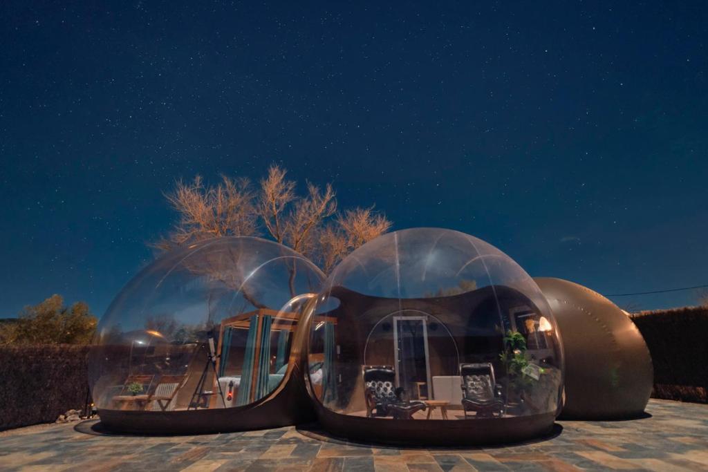 Burbuja de hotel iluminada de noche bajo un cielo estrellado, con zona de estar interior visible a través del domo transparente