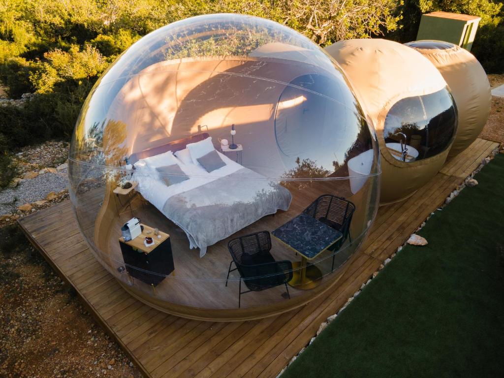 Cúpula de hotel burbuja con cama doble, zona de desayuno con mesa y sillas, ubicada sobre plataforma de madera en entorno natural