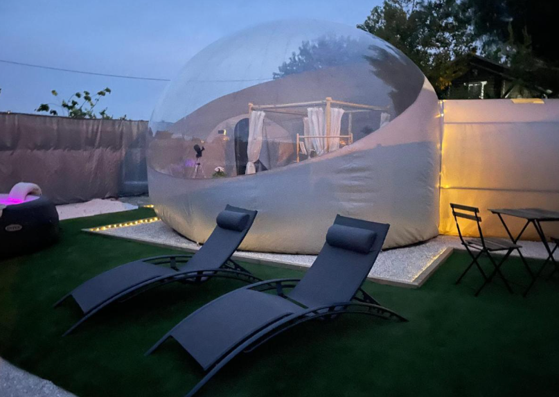 Burbuja de hotel iluminada al anochecer con cama visible en el interior, tumbonas en el jardín y zona de estar exterior