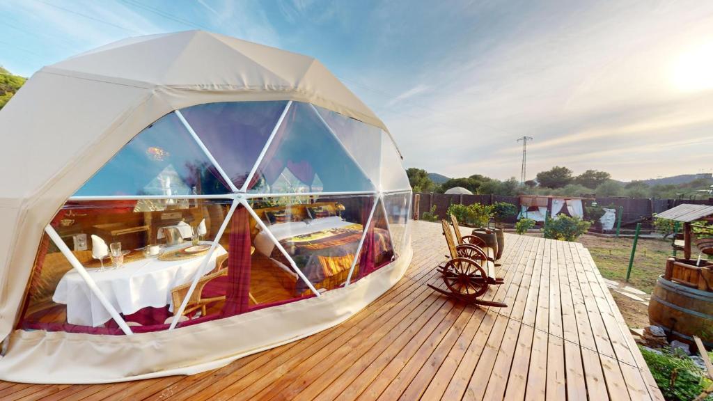 Hotel burbuja con fachada transparente, interior elegante con mesa puesta y cama, ubicado sobre una terraza de madera con vistas al paisaje natural al atardecer