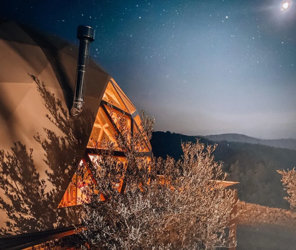 Hotel burbuja iluminado al anochecer entre árboles, con cielo estrellado y luna brillante al fondo