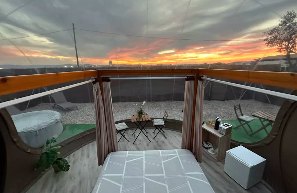 Vista desde el interior de una burbuja de hotel al atardecer, con cama, mesa para dos, cafetera, minibar y bañera exterior visible al fondo