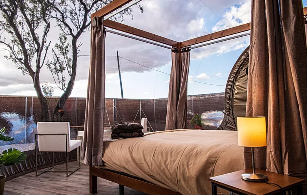 Interior de burbuja de hotel con cama con dosel, lámpara encendida, sillas blancas y vistas al exterior a través de la estructura transparente