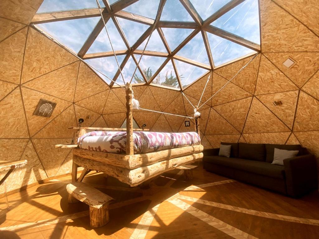Interior de una cabaña con cama colgante de madera, techo transparente con vista al cielo y sofá oscuro en una esquina