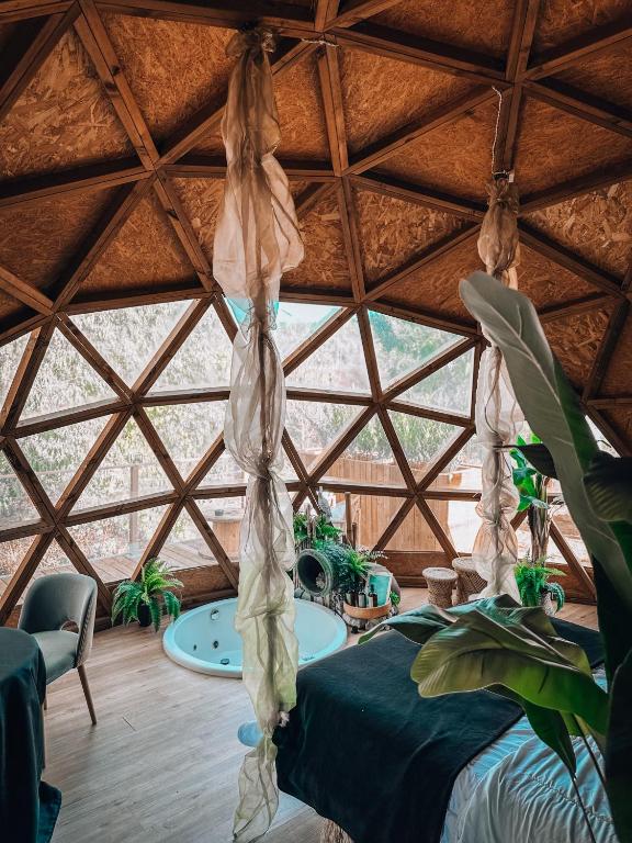 Interior de una cúpula geodésica de madera con jacuzzi, cama, sillas y plantas decorativas, iluminado con luz natural a través de ventanas triangulares
