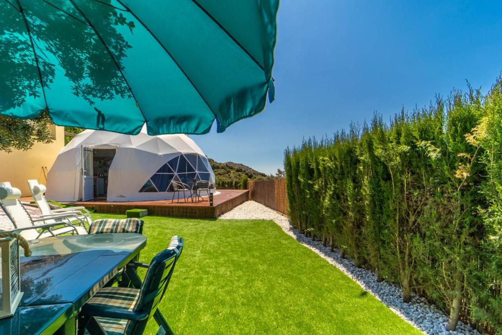Domo geodésico blanco con terraza de madera y sillas al aire libre, rodeado de césped, setos altos y una sombrilla verde.