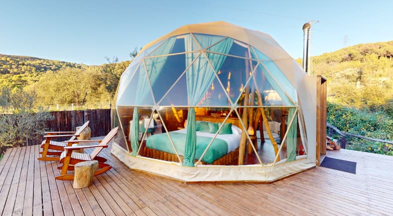 Hotel burbuja de estructura geodésica con cama grande, cortinas turquesa, terraza de madera y rodeado de naturaleza